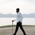 Stylish man in white jacket walking on golf course at sunset by a lake.