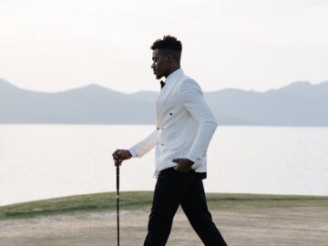Stylish man in white jacket walking on golf course at sunset by a lake.