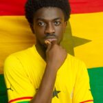 Young man posing in front of Ghana flag, wearing a yellow sports jersey.