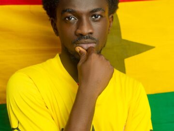 Young man posing in front of Ghana flag, wearing a yellow sports jersey.