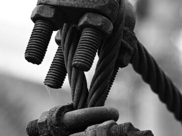 Black and white close-up of a cable tension system with bolts and rust detailing.