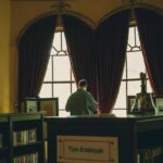 A man stands browsing books in a sophisticated library with ornate curtains and chandeliers.