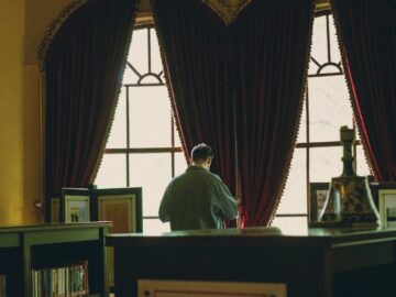 A man stands browsing books in a sophisticated library with ornate curtains and chandeliers.