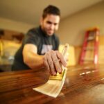 A man carefully applies varnish to a wooden surface indoors, using a paintbrush for a smooth finish.
