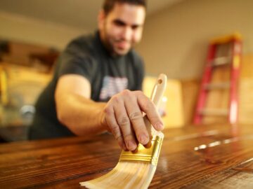 A man carefully applies varnish to a wooden surface indoors, using a paintbrush for a smooth finish.