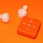 Close-up of wireless earphones and a compact music player on an orange background, showcasing modern audio technology.