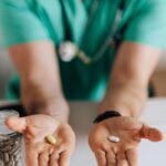 Crop man doctor wearing medical uniform demonstrating medicine to patient in clinic during work sitting at table with stethoscope and clipboard