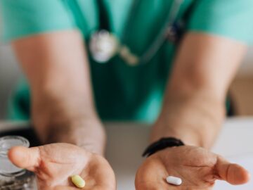 Crop man doctor wearing medical uniform demonstrating medicine to patient in clinic during work sitting at table with stethoscope and clipboard