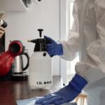 Person in protective suit cleaning a kitchen counter with spray bottle and gloves.