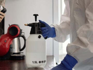 Person in protective suit cleaning a kitchen counter with spray bottle and gloves.