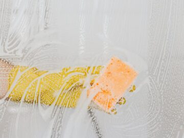 A gloved hand cleans glass with a sponge and soapy water, ensuring hygiene.