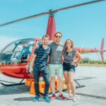Three friends smiling in front of a red helicopter at the airfield, enjoying a sunny day.