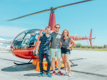 Three friends smiling in front of a red helicopter at the airfield, enjoying a sunny day.