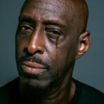 A close-up portrait of a man shedding a tear against a gray background, depicting sadness and contemplation.