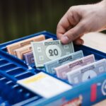 Crop unrecognizable person showing game banknote from collection in box while playing Monopoly at table