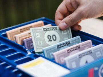 Crop unrecognizable person showing game banknote from collection in box while playing Monopoly at table