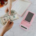 Close-up of hands exchanging US dollars with a pink calculator on a marble surface.