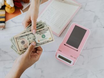 Close-up of hands exchanging US dollars with a pink calculator on a marble surface.