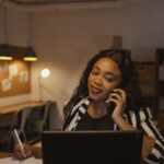 Confident businesswoman multitasking on phone and laptop in a warm office atmosphere.
