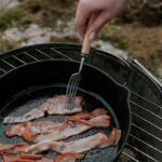 Close-up of bacon strips frying in a pan on a charcoal grill outdoors.