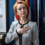 Professional woman in office attire showing respect with hand on chest and American flag in background.