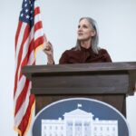 A woman speaking at a podium with the American flag, symbolizing leadership and patriotism.