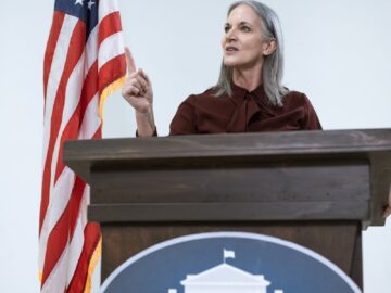A woman speaking at a podium with the American flag, symbolizing leadership and patriotism.