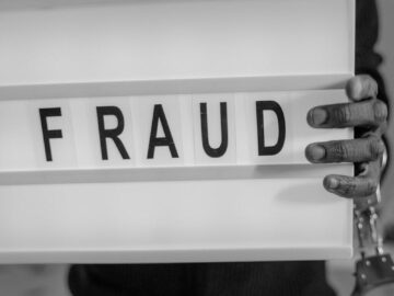 Grayscale photograph of a handcuffed individual holding a letter board with the word 'FRAUD'.