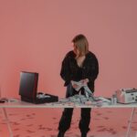 A woman standing at a desk counting cash with a briefcase and money counter in a red-lit room.