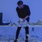 A man interacts with scattered dollar bills in a blue-lit modern setting.