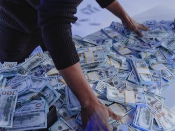 Adult man organizing a massive pile of US dollar bills indoors in dynamic lighting.