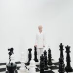 A person in a white suit stands among giant chess pieces on a board, symbolizing strategy.