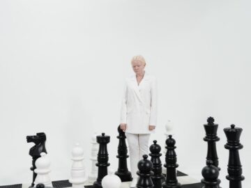 A person in a white suit stands among giant chess pieces on a board, symbolizing strategy.