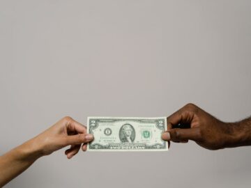A dollar bill is exchanged between two diverse hands on a neutral background.