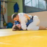 Two martial artists practicing judo grips and techniques on a yellow dojo mat indoors.