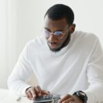 Man in glasses working on calculations at desk in bright office setting.