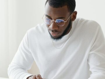Man in glasses working on calculations at desk in bright office setting.