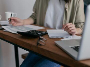 A woman manages finances at home, using a laptop and calculator on a wooden desk.
