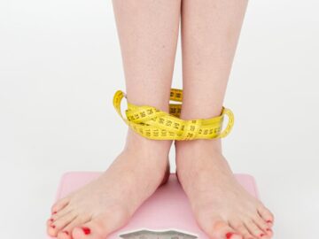 High angle of crop anonymous female with centimeter on legs checking weight on scales on white background