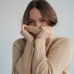 Young content female covering face with warm beige sweater and looking at camera in light studio