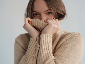 Young content female covering face with warm beige sweater and looking at camera in light studio