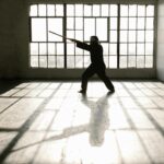Silhouette of martial artist practicing with a staff in a sunlit studio.
