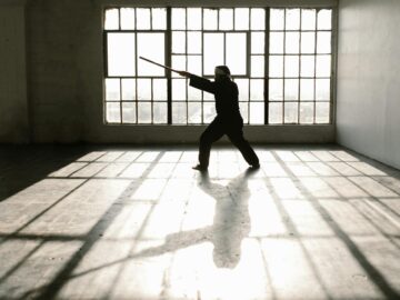 Silhouette of martial artist practicing with a staff in a sunlit studio.