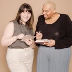 Two women, one holding a notebook and pen, smiling while collaborating together indoors.