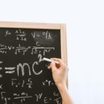 Close-up of hand writing physics equations on a chalkboard indoors.