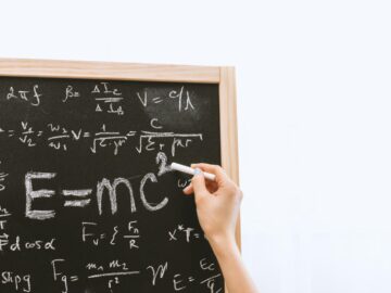 Close-up of hand writing physics equations on a chalkboard indoors.