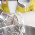 Crop unrecognizable female confectioner pouring melted sugar syrup into planetary mixer with whipped egg whites