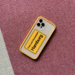 Smartphone with vibrant yellow case displaying 'Hamburg' on a textured pink carpet.