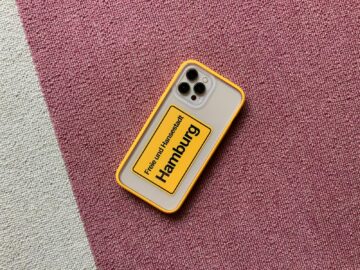 Smartphone with vibrant yellow case displaying 'Hamburg' on a textured pink carpet.