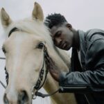 A man wearing a black leather jacket gently embraces and leans against a horse.